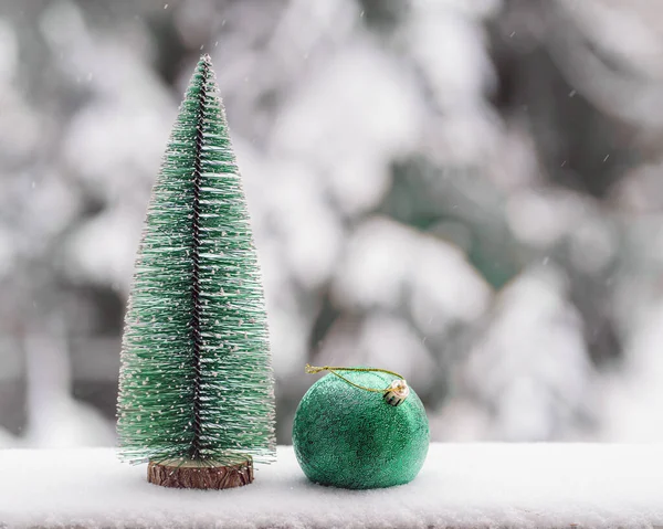 Oyuncakları olmayan yeşil bir Noel ağacı, kar yağışı sırasında bulanık yeşil çam ağaçlarının doğal arka planında, doğal karın üzerinde yuvarlak yeşil bir oyuncak. Minimalist Noel şablonu. Boşluğu kopyala