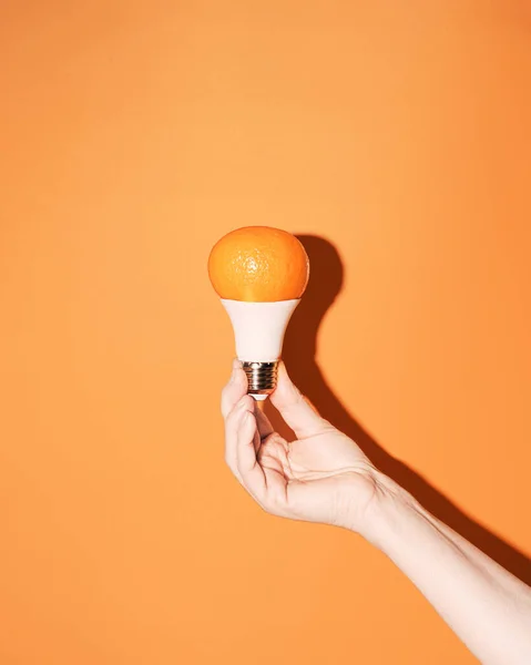 Minimalist creative concept of renewable energy, respect for natural resources, sustainable development of technology in unity with nature. Led lamp with tangerine dome in female hand