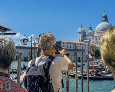 A real traveler with a backpack on his back takes a smartphone photo of the seascape with gondolas and historic buildings in the Gulf of Venice. Tourist journeys through authentic European cities