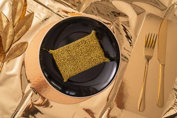 Ironic gold food and hygiene concept in a luxurious art deco style, gold metal kitchen sponge on a black plate, gold knife and fork and a gold branch with leaves lie next to it on a gold tablecloth