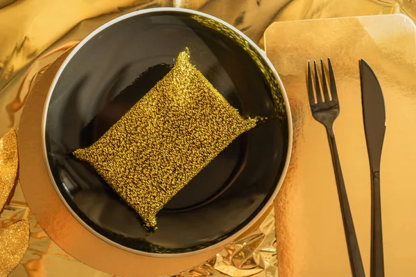 Gold food and hygiene concept in luxurious art deco style, ray of sun on a gold metal kitchen sponge on a black plate, black knife and fork next to each other on a gold tablecloth, gold branch