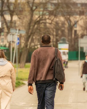 Süet ceketli siyah bir adamın arkası, elinde telefon olan bir kot pantolon ve bir bahar günü yoldan geçenler arasında omzunda yumuşak bir çanta.