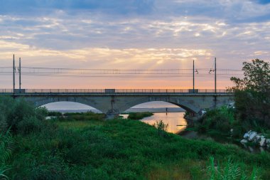 Nehrin ağzındaki demiryolu köprüsü şafak vakti denize dökülüyor, güneş ışınları bulutlarda parlıyor, nehrin ve denizin yüzeyini yansıtıyor, balıkçı balık tutuyor.
