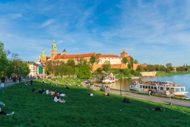 Krakow, Polonya - Nisan 2023: Wawel kraliyet kalesi bir tepenin üstünde bir su kütlesinin yanında, insanlar arka planda bir ortaçağ şatosunun eteğinde yeşil çimlerin üzerinde dinleniyorlar, güzel şehir manzarası, eski şehir