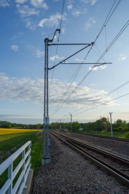 Güneşli bir akşamda, üzerinde elektrik kabloları olan demiryolu hattı, yüksek metal direkler, temiz gökyüzü ve doğal yeşil zemin, güzel bir bahar doğasına karşı demiryolu, demiryolu seyahati