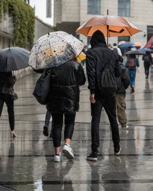 Kötü hava koşullarında yoldan geçenler şemsiyeli bir caddede yağmurda yürürler, yağmurda bir tarih, ıslak bir yolda ıslanmış yolcuların yansımaları, yağmur damlaları ve su birikintileri. Dikey mahsul