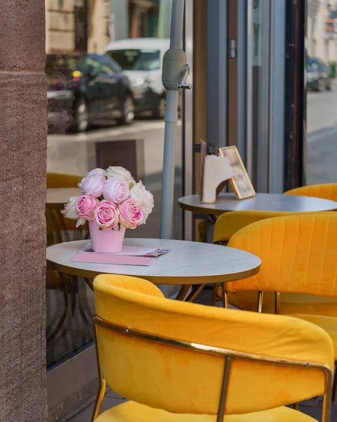 Bouquet of pink roses on a table of a street cafe against the background of reflections of parked cars in a glass window, yellow chairs next to tables on a city street, artificial flowers