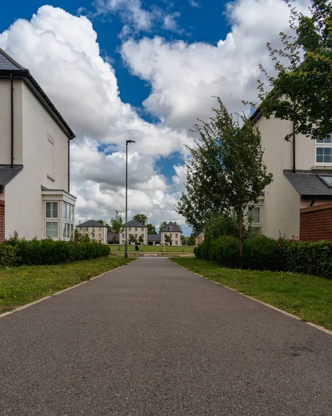 İngiliz kırsalında, çiftlik evlerine ve yeşil çimenlere giden asfalt bir yolun fotoğrafı. Gökyüzü bulutlu ve yağmurlu. Asfalt dokusu ve doğrusal perspektif derinlik ve karşıtlık ekler