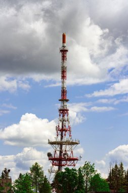 Beyaz bulutlu mavi gökyüzüne karşı uzun kırmızı ve beyaz bir telekomünikasyon kulesi. Kulede antenli ve çanaklı bir kafes yapısı var. Tepesinde ağaçlarla çevrili bir deniz feneri var.