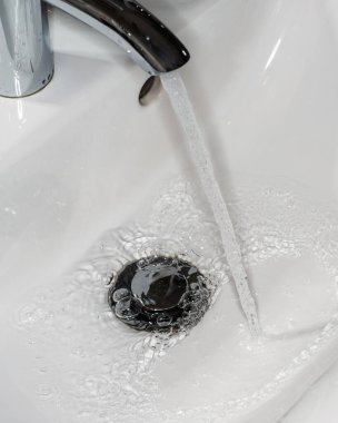 Parlak metal bir musluk ve beyaz bir lavabonun üzerinde bir su akıntısı. Hijyen ve iç tasarım kavramları için minimalist ve zarif bir imaj. Dikey çerçeve. Yakın plan.
