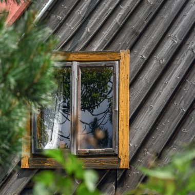 Köy evinin eğimli çatısında, ağaçların ve gökyüzünün dingin güzelliğini yansıtan eski bir ahşap pencere. Ebedi cazibe ve doğanın kucaklaması kavramını yakalamak.