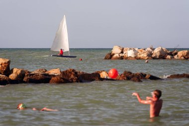 Yelkenli ve yüzen çocuklarla dolu sakin bir deniz manzarası, kayalar ve şamandıralarla işaretlenmiş, boş zamanların ve seyahatlerin özünü yakalıyor.