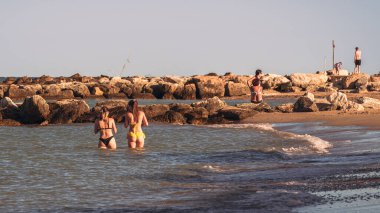 Huzurlu bir sahilde altın bir saat, kayalık kıyılarda eğlenen insanlar, dalgalar yavaşça çarpıyor. Bir huzur ve rahatlama anı.