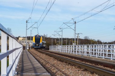 Elektrikli tren raylarında modern sarı tren. Tepemizde elektrik hatları ve beyaz güvenlik bariyerleri var. Demiryolu altyapısı toplu taşımayı ve sürdürülebilir seyahat teknolojisini gösteriyor