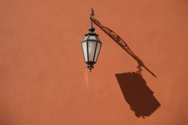Eski bir fener, güneş ışığında turuncu bir duvara gölge düşürür. Bu klasik sokak lambası nostaljik ve romantik bir atmosfer yaratır. Görüntü mimariyle ilgili projeler için mükemmel.