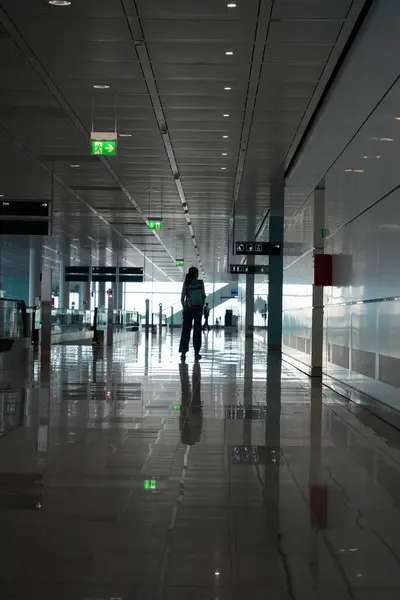 Bir gezgin sabahın erken saatlerinde geniş bir havaalanı koridorunda, yansıtıcı yüzeyler ve aydınlatma işaretleriyle çevrili, beklentinin özü ve modern seyahat, yaşam tarzı görüntüsü için ideal.