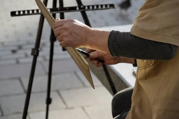 Sanatçının eli tuvale dikkatlice çizim yapıyor, açık havada hareketli bir ortamda yaratıcılığın özünü yakalıyor.