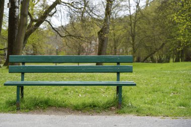 Boş yeşil park bankı bahar parkındaki patikanın yakınındaki çimlerde huzur içinde dinleniyor. Rahatlama kavramını, doğada yalnızlığı ya da derin düşüncelere dalma anını temsil eder.