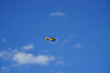 Uçsuz bucaksız mavi gökyüzünde uçan sarı helikopter. Beyaz bulutlar modern teknoloji ve doğanın kesişimini temsil ediyor. Havacılık taşımacılığı ve yan yana gelme kavramı