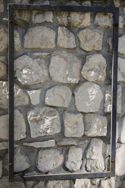 Düzensiz bej, gri taşlar ve koyu harç eklemleri olan taştan bir duvarın doku arka planını kapat. Boş metal çerçeve detayı geometrik kontrastı ekler. Mimari yüzey fotoğrafı