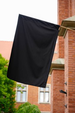 Gotik tarzı kemerleri ve yeşil yaprakları olan tarihi kırmızı tuğlalı bir binaya karşı direkte asılı siyah bir bayrak resmi veya sembolik bir görüntü.