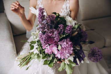 Wedding. Young bride in a white dress sits on the couch holding a bouquet of pink and purple flowers with greenery