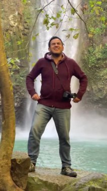 Erkek fotoğrafçı kollarını muzaffer bir şekilde kaldırıyor ve arkasında bir şelale var.