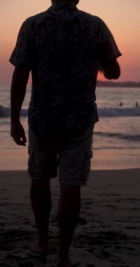 Adamın sihoueti sahilde gün batımına doğru yürüyor, duruyor ve uzaklaşıyor.