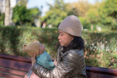 Güneşli bir günde şehir parkında şefkatli ve tatlı bebeğini kucağında tutan küçük sevimli bir kızın annesi olarak genç ve güzel Asyalı bir kadının açık hava yaşam tarzı portresi.