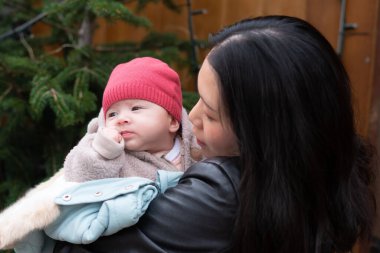 Genç, mutlu ve çekici Asyalı Çinli bir kadın Noel ağacının önünde küçük kızı kollarında tutuyor ve anne-kız kavramı ile kış marketinde yürüyor.