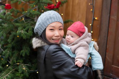 Genç, mutlu ve çekici Asyalı Çinli bir kadın Noel ağacının önünde küçük kızı kollarında tutuyor ve anne-kız kavramı ile kış marketinde yürüyor.