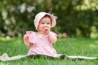 Parktaki sevimli ve mutlu bir kız bebek. 7-8 aylık güzel bir çocuğun portresi. Şehir parkında çimlerin üzerinde oturmuş, rahat bir şapka ve elbiseyle gülümsüyor.