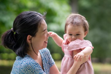 Anne ve kızının açık hava yaşam tarzı portresi. Genç, mutlu ve tatlı Asyalı Koreli kadın 8 aylık bebeğiyle şehir parkında oynuyor.