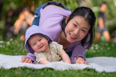 Şehir parkında 5 aylık mutlu ve sevimli bir kız bebek ve onun Asyalı Koreli kadını, sevgi ve anne-kız arasındaki yemyeşil çimlerin arasında bir battaniyede neşeli bir anı paylaşıyorlar.
