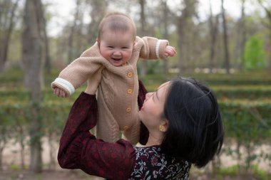 Genç, mutlu ve güzel Asyalı bir kadın kucağında küçük bir kız çocuğu, sevimli bir kız bebek, parkta birlikte oynaşıyorlar anne ve çocuk sevgisi ve ebeveynlik kavramında.