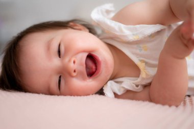 9 aylık karışık etnik kökenli Asyalı beyaz kız bebek portresi. Mutlu ve tasasız oynuyor. Yatakta emekliyor, gülüyor ve çocukluk ve çocuk odası konseptinde heyecanlanıyor.