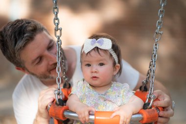 Açık hava yaşam tarzı, babanın oyun parkında neşeli gülümseyen güzel ve tapılası bir bebekle oynadığı, çocukluk konseptinde salıncakta şirin bir kurdele taktığı bir resim.