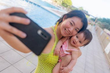 Asyalı bir kadın, sevimli küçük kızıyla fotoğraf çekiyor. Güzel Koreli bir anne, kızını kucağında tutuyor ve tatillerde cep telefonuyla fotoğraf çekiyor.