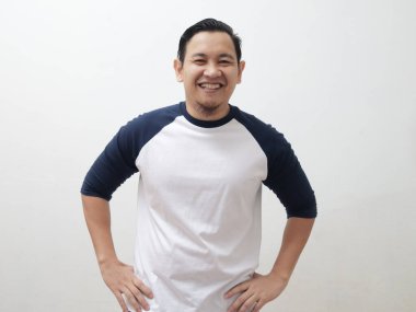 Beyaz arka planda dikilirken elleri kalçalarında gülümseyen sıradan bir tişört giyen çekici Asyalı adam.