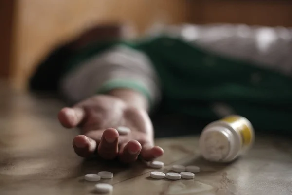 Anonymous man lying on the floor, unconscious or dead due to drugs abuse, focus on fingers with pills