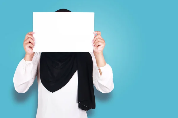 Portrait of muslim woman in hijab covering her face with blank empty paper. copy space mock up for anonymouse person, over blue background