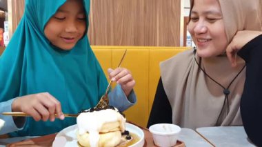 Asyalı Müslüman kadın kahvaltısını yapıyor ya da kızıyla kafede öğle yemeği yiyor, insanlar yemek yiyor, mutlu ebeveyn ve çocuk bağı kuruyor.