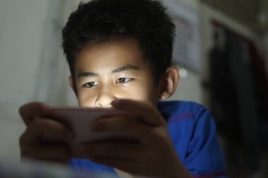 Küçük Asyalı çocuk internetten izliyor ya da akıllı telefonuyla oyun oynuyor. Akşamları yatakta. Cihaz bağımlılığı olan çocuğu kapatıyor.