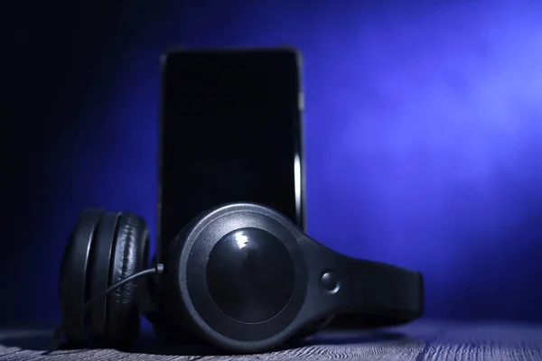 ヘッドフォンとブラックスクリーンスマートフォン 暗闇の青色光の下で 音楽やポッドキャストアプリのコンセプトを聞く — ストック写真
