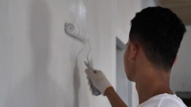 Asyalı erkek ressam paten fırçasıyla yeni bir beyaz duvar boyuyor, ev dekorasyonu konsepti.