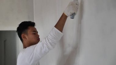 Asyalı erkek ressam paten fırçasıyla yeni bir beyaz duvar boyuyor, ev dekorasyonu konsepti.
