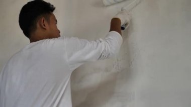 Asyalı erkek ressam paten fırçasıyla yeni bir beyaz duvar boyuyor, ev dekorasyonu konsepti.