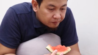 Asyalı adam telefonuyla izlerken karpuz meyvesi yiyor, sağlıklı atıştırma zamanı.