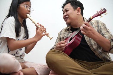 Asyalı bir adam kızına gitar çalarken flüt çalmayı öğretiyor. Müzik öğretmeni ve öğrenci birlikte oynuyorlar.