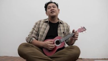 Asyalı adam evde oturmuş gitar çalarken şarkı söylüyor.